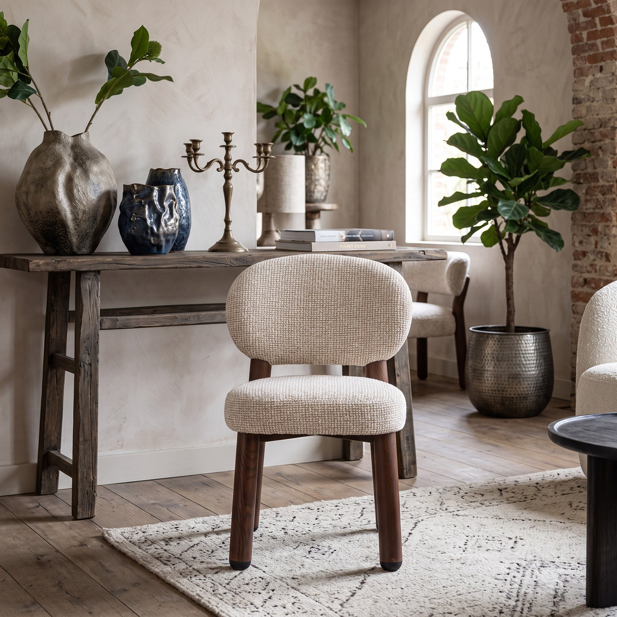 Vooraanzicht van de sculpturale Oatly eetkamerstoel op een geweven vloerkleed, omringd door een houten sidetable met aardewerk vazen, kandelaar en een grote plant in een bronzen pot.