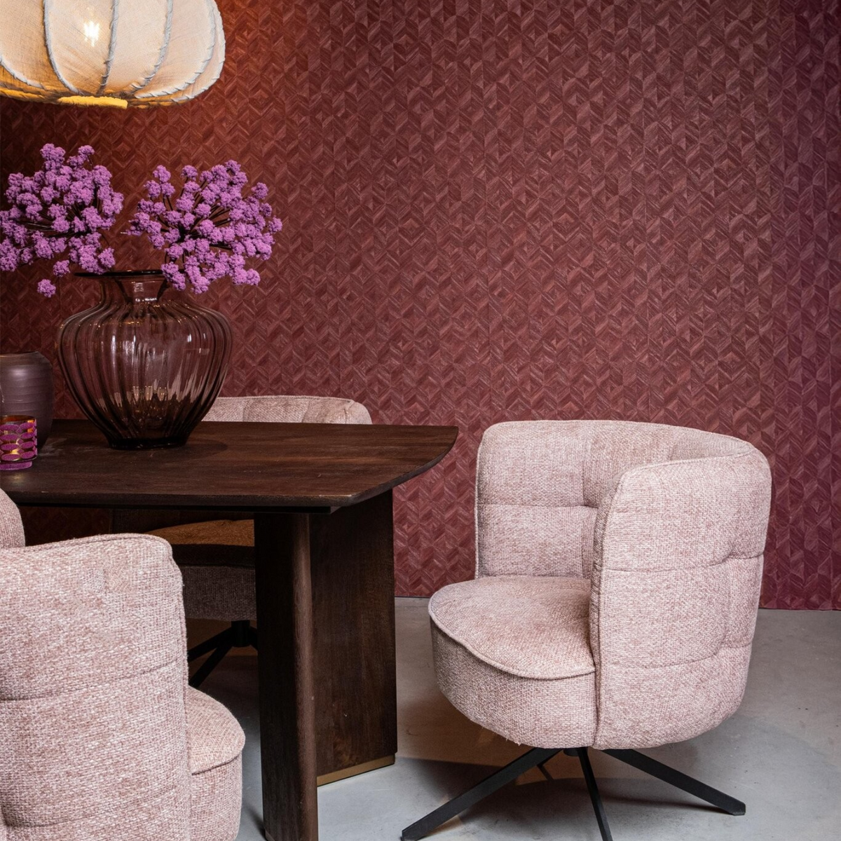Een sfeervolle hotel-chique eethoeksetting met de zachtroze draaistoel aan een donkere eettafel, gedecoreerd met een glazen vaas met paarse bloemen en een warme bordeauxrode wandafwerking.