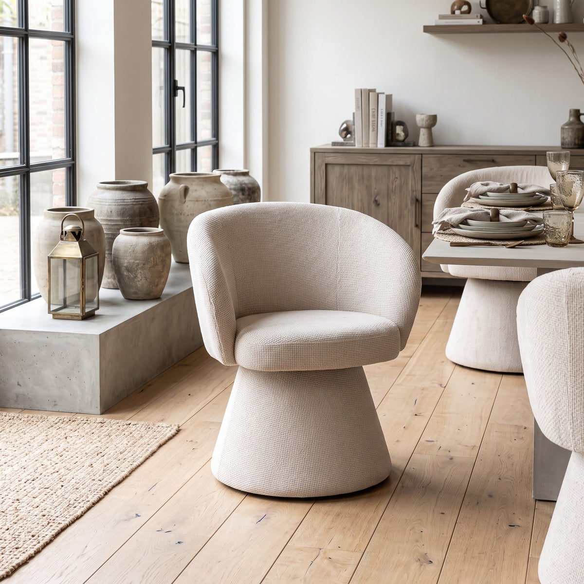 Een rustgevende eethoeksetting met de crèmekleurige fauteuil op een houten vloer, omringd door een lichte eettafel, keramiek en robuuste aardewerk potten bij grote raampartijen.