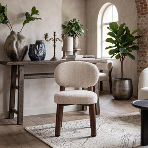 Vooraanzicht van de sculpturale Oatly eetkamerstoel op een geweven vloerkleed, omringd door een houten sidetable met aardewerk vazen, kandelaar en een grote plant in een bronzen pot.