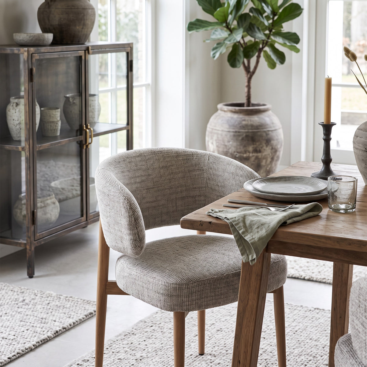 De comfortabele armstoel Okura, sfeervol opgesteld aan een robuuste houten eettafel in een lichte eethoek. Op de achtergrond een vintage metalen vitrinekast en een grote aardewerk pot met plant, voor een serene en uitnodigende sfeer.
