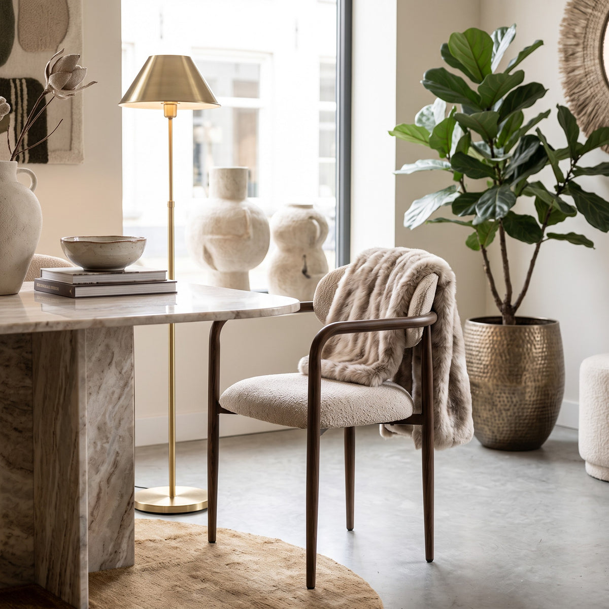De elegante armstoel met houtlook frame en zachte tan bekleding, sfeervol gestyled aan een royale marmeren eettafel met een imitatiebont plaid, gouden design vloerlamp en grote kamerplant in bronzen pot.