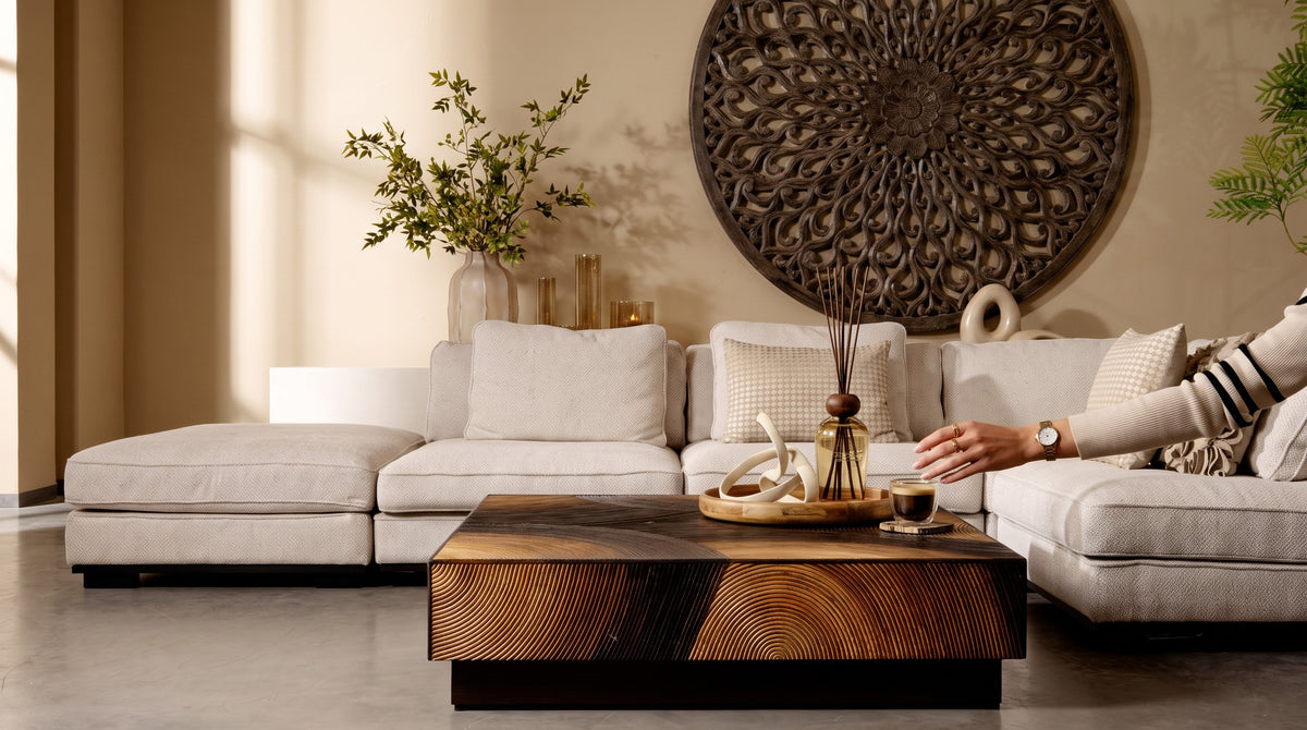 Een sfeervol collectie-overzicht van een luxe woonkamer met een grote lichte hoekbank, een opvallende houten salontafel met uitgesneden motief, gedecoreerd met geurstokjes en een kopje koffie, en een grote ronde houten wanddecoratie van PTMD.