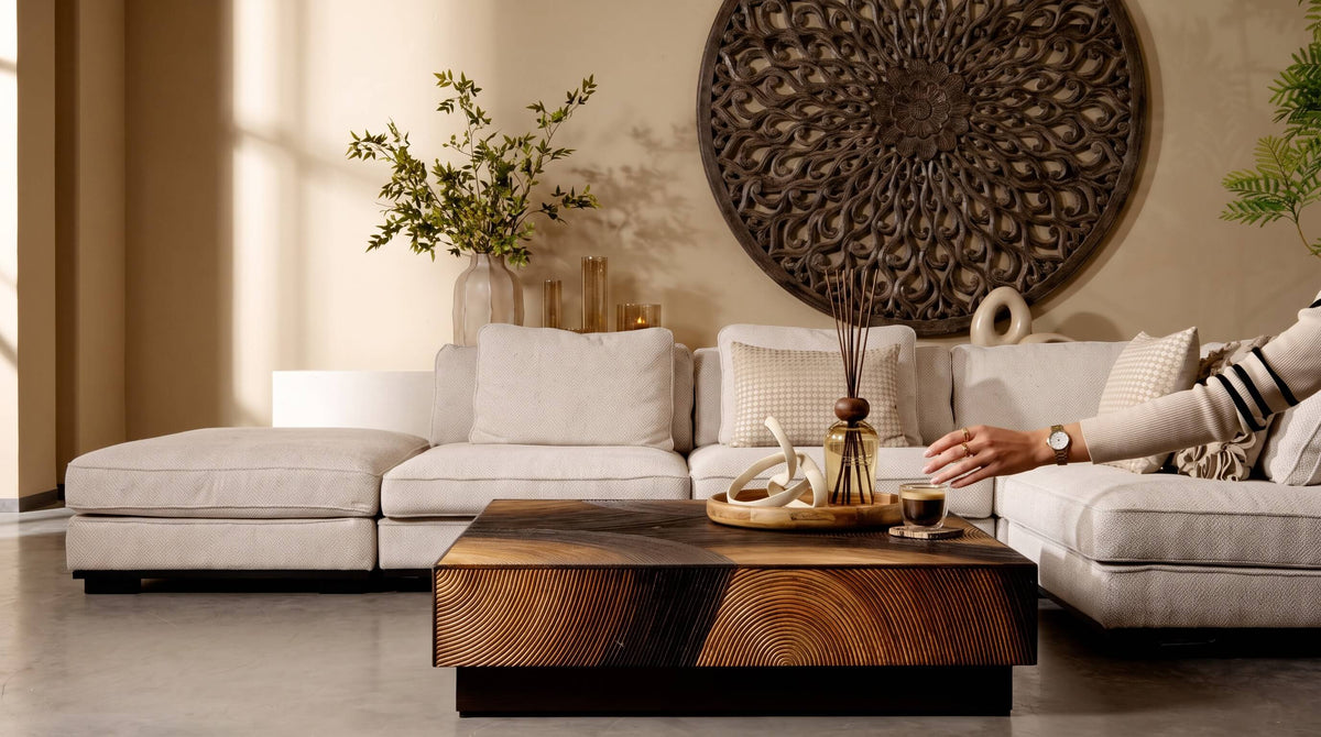Een sfeervol collectie-overzicht van een luxe woonkamer met een grote lichte hoekbank, een opvallende houten salontafel met uitgesneden motief, gedecoreerd met geurstokjes en een kopje koffie, en een grote ronde houten wanddecoratie van PTMD.