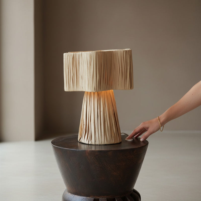 Een natuurlijke raffia tafellamp op een donkerhouten krukje waarbij een hand de schakelaar aanraakt, dienend als navigatielink naar de verlichting collectie met subcategorieën zoals hanglampen, tafellampen en vloerlampen.